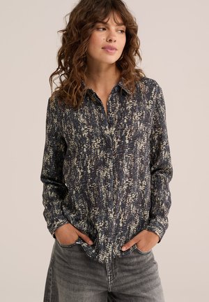 Femme aux cheveux bouclés portant une chemise à manches longues foncée et à motifs et un jean gris, debout les mains dans les poches, regardant légèrement sur le côté.