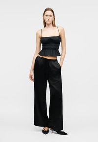 Γυναίκα που φοράει μαύρο αμάνικο τοπ με βολάν, μαύρο παντελόνι φαρδιάς γραμμής και μαύρες μυτερές γόβες, στέκεται με το ένα χέρι στην τσέπη.