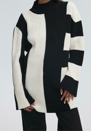 Pull en maille côtelée noir et blanc avec col montant, manches longues évasées et blocs de couleurs audacieux verticaux et horizontaux porté par une personne.