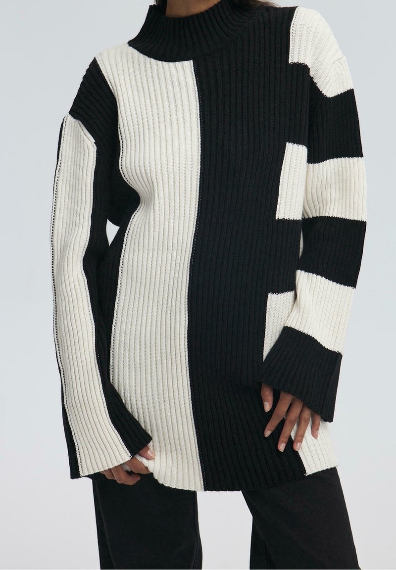 Pull en maille côtelée noir et blanc avec col montant, manches longues évasées et blocs de couleurs audacieux verticaux et horizontaux porté par une personne.