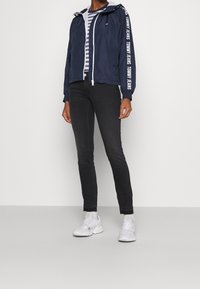 Chaqueta cortavientos de color navy con capucha, cremallera blanca y mangas con marca. Combinada con jeans skinny negros y zapatillas blancas; debajo, una camiseta a rayas.