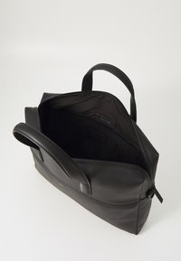 Schwarze Lederhandtasche mit glatter Textur, Reißverschluss, zwei Griffen und einem offenen Innenraum, der mit einem Stofffutter und einem Markenetikett ausgestattet ist.