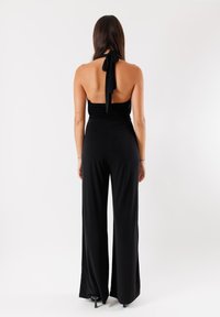 Jumpsuit nero con scollo all'americana, corpino smockato, schiena scoperta, gambe ampie e dettaglio con fiocco al collo, realizzato in un tessuto morbido ed elastico.