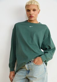 Žalias džemperis ilgomis rankovėmis ir apykaklės modeliu, su maža balta Nike logotipu ant krūtinės. Pora su šviesiai mėlynomis džinsais.