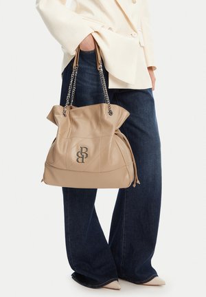 Person trägt weite, dunkle Jeans und cremefarbenen Blazer, hält beigefarbenen gesteppte Lederhandtasche mit Kettenriemen und Metalllogo.