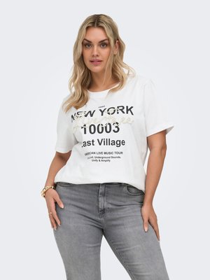 Blondynka w białej koszulce z grafiką Nowego Jorku i szarych dżinsach, stojąca z rękami na biodrach na jasnoszarym tle.