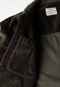 Ciemnooliwkowa kurtka z korduroy w pionowe prążki, z zamkiem błyskawicznym i jedną kieszenią. Metka wewnątrz mówi "SANGAR."