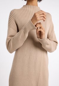 Beige gestricktes Kleid mit hohem Kragen, das eine Mischung aus vertikalen und horizontalen Mustern aufweist, lange Ärmel und eng anliegende Bündchen hat.
