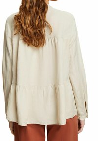 Blusa larga de manga larga beige con diseño de peplum. Fabricada en tela ligera, cuenta con un yugo trasero y puños abotonados.