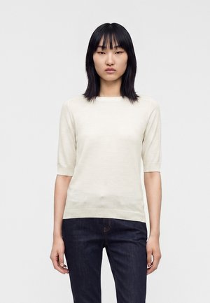 Donna con capelli neri lisci che indossa un maglione bianco a maniche corte e jeans blu scuro, in piedi davanti a uno sfondo semplice.