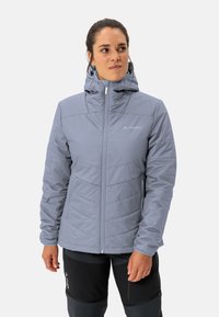 Giacca isolante grigia chiara con cappuccio, frontale con zip e texture trapuntata. Logo sul petto e polsini elasticizzati. Indossata con pantaloni scuri.