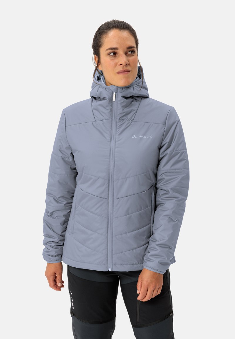 Giacca isolante grigia chiara con cappuccio, frontale con zip e texture trapuntata. Logo sul petto e polsini elasticizzati. Indossata con pantaloni scuri.