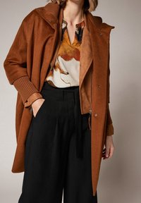 Abrigo marrón de gran tamaño con un acabado texturizado, que cuenta con un cierre frontal con cremallera, capucha y puños acanalados. Lleva puesto sobre una blusa de estampado multicolor.