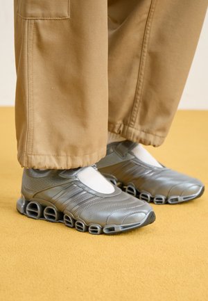 Baskets de sport argentées avec un design épuré, un matériau synthétique texturé et une semelle découpée distinctive. Portées avec un pantalon beige et des chaussettes blanches.