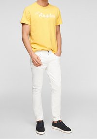 Uomo che indossa una maglietta gialla con la scritta "Los Angeles", jeans bianchi arrotolati e sneakers blu navy su uno sfondo leggero e neutro.