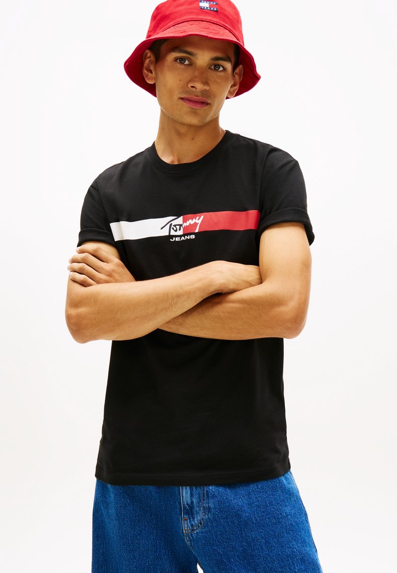 Νέος άνδρας με μαύρο t-shirt Tommy Jeans, κόκκινο καπέλο τύπου bucket και μπλε τζιν, στέκεται με σταυρωμένα τα χέρια σε λευκό φόντο.