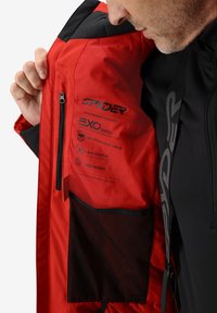 Intérieur de la veste rouge avec des accents noirs, étiquettes Exo Shield pour l'imperméabilité, tissu extensible dans les quatre sens, et une poche en mesh sur le côté gauche.