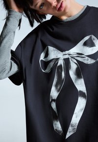 Sort t-shirt med et stort, hvidt buegrafik. Lange grå ærmer er synlige. Stoffet ser blødt ud med en afslappet pasform.