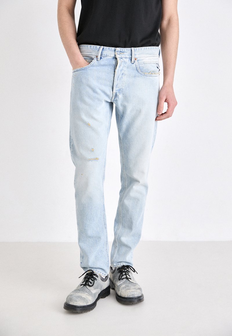Jean délavé bleu clair avec détails déchirés, coupe slim, comportant cinq poches et une taille standard. Chaussures noires avec une finition texturée.