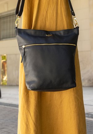 Crna kožna torba sa teksturom na ramenu, sa zlatnim zatvaračem i metalnim detaljima, nošena preko haljine boje gorušice u urbanom okolišu na otvorenom.