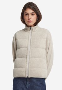 Ljusbeige zip-jacka med ribbade kragar, med vadderade sektioner och en mjuk textur. Framdels dragkedjestängning, avslappnad passform.