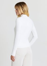 Weißes Langarmoberteil mit Stehkragen, gerippter Textur und figurbetontem Schnitt. Kombiniert mit cremefarbenen Cordhosen mit Gesäßtaschen.