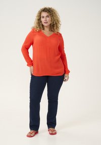 Rote Bluse mit langen Ärmeln und V-Ausschnitt, aus leichtem Material, kombiniert mit dunkelblauen Jeans und roten Flip-Flops vor einem neutralen Hintergrund.