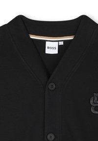 Cardigan en coton noir tricoté avec un décolleté en V profond, deux boutons et un détail de logo texturé sur le côté gauche. Finition intérieure contrastante en or et blanc.