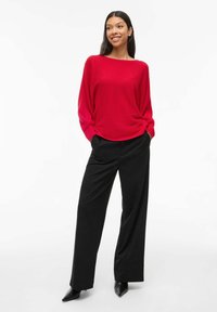 Pull rouge à côtes avec des manches longues associé à un pantalon large noir taille haute et des talons aiguilles noirs, sur fond blanc.