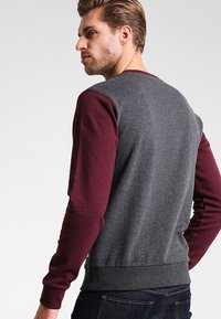 Sudadera gris con mangas burdeos, cuello redondo y dobladillo acanalado. La tela parece suave con una textura lisa y un ajuste casual.