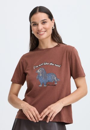 Kvinde smiler, iført en brun t-shirt med en gravhund-grafik og teksten "I'm not like the rest. Born to be cute" foran.