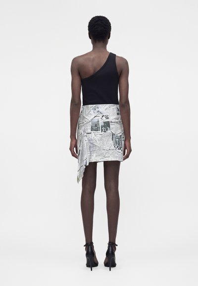 Γυναίκα που στέκεται με την πλάτη, φοράει ένα μαύρο τοπ με έναν ώμο και ασύμμετρη φούστα με σχέδιο από κείμενα και εικόνες εφημερίδων.