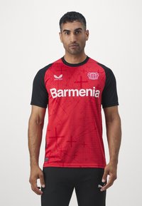 Camisola desportiva vermelha com mangas pretas, apresentando um padrão geométrico, logótipo e texto em branco em negrito. Feita de um tecido leve e respirável.