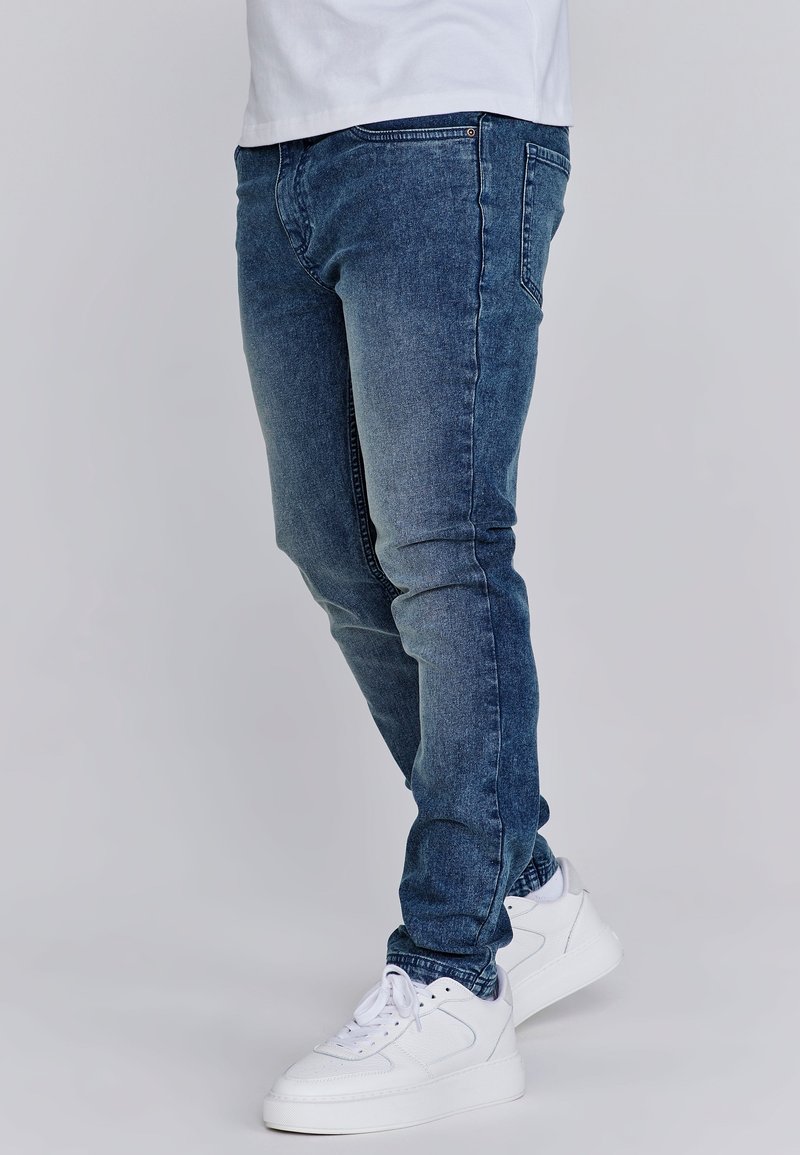 Personne portant un jean bleu ajusté et des baskets blanches, debout contre un fond gris clair uni.