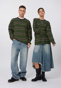 Grün gestreifter Strickpullover, hellblaue weite Jeans und ein Jeans-Midirock mit fransigem Saum; schwarze Knöchelstiefel mit Schnallen-Detail.
