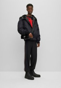 Chaqueta negra acolchada con capucha forrada de pelo sintético, suéter de rayas rojas y negras, pantalones deportivos negros con rayas laterales y botas negras gruesas.