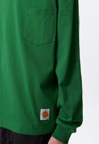 Green long-sleeve shirt made of cotton, featuring a left chest pocket and a logo patch at the hem. Smooth texture and relaxed fit.