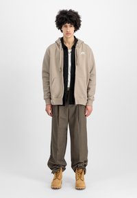 Sudadera beis con cremallera y cordones, camisa negra de botones, pantalones grises y botas marrones con cordones amarillos. Diseño minimalista, ajuste casual.