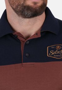 Polo à manches courtes en bleu marine et rouille, présentant un design bicolore, un col à trois boutons et un logo brodé sur la poitrine.