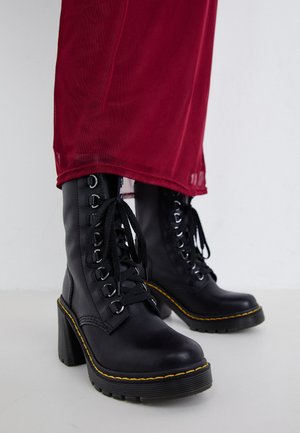 Black leather ankle boots with a chunky heel, featuring lace-up design, silver eyelets, and yellow stitching along the sole.