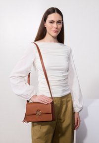 Borsa a tracolla marrone con superficie testurizzata, dettagli in hardware dorato e accentuazione con frange. Indossata con un top bianco a maniche lunghe arricciato e pantaloni oliva.