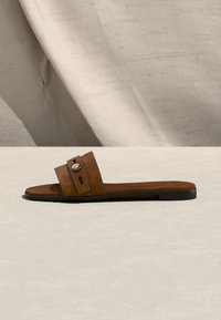 Sandale mule en cuir marron avec deux brides, détail bouton en métal, et semelle plate noire sur une surface claire et texturée.