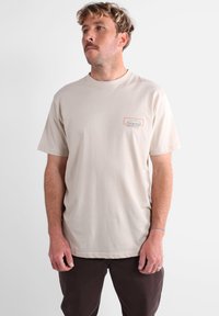 T-shirt en coton beige avec un col rond, des manches courtes et une petite écusson logo orange et bleu sur la partie gauche de la poitrine.