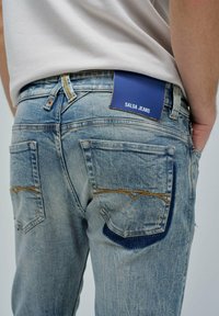 Vaqueros de mezclilla azul claro con un lavado desgastado, que presentan dos bolsillos traseros y una etiqueta azul marino con la inscripción "SALSA JEANS". Costuras visibles que acentúan los bolsillos.