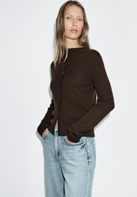 Brown ribbed cardigan with a high neckline, button front, and long sleeves, paired with light blue high-waisted jeans. Soft texture.