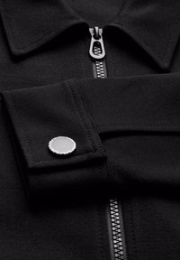 Detail einer schwarzen Stoffjacke mit silbernem Reißverschluss und Knopf an den Manschetten, umgeben von weißem Text am Rand. Zu sehen sind der Kragen und der gefaltete Ärmel.