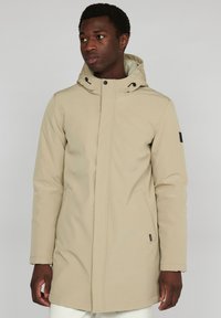 Parka de color beig, impermeable, con capucha, cierre frontal de cremallera, botones a presión, bolsillos laterales y un detalle de etiqueta negra en la manga izquierda.