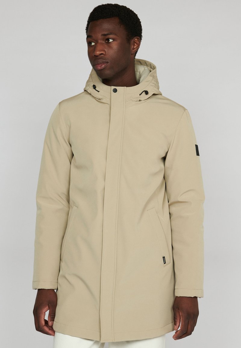 Parka de color beig, impermeable, con capucha, cierre frontal de cremallera, botones a presión, bolsillos laterales y un detalle de etiqueta negra en la manga izquierda.