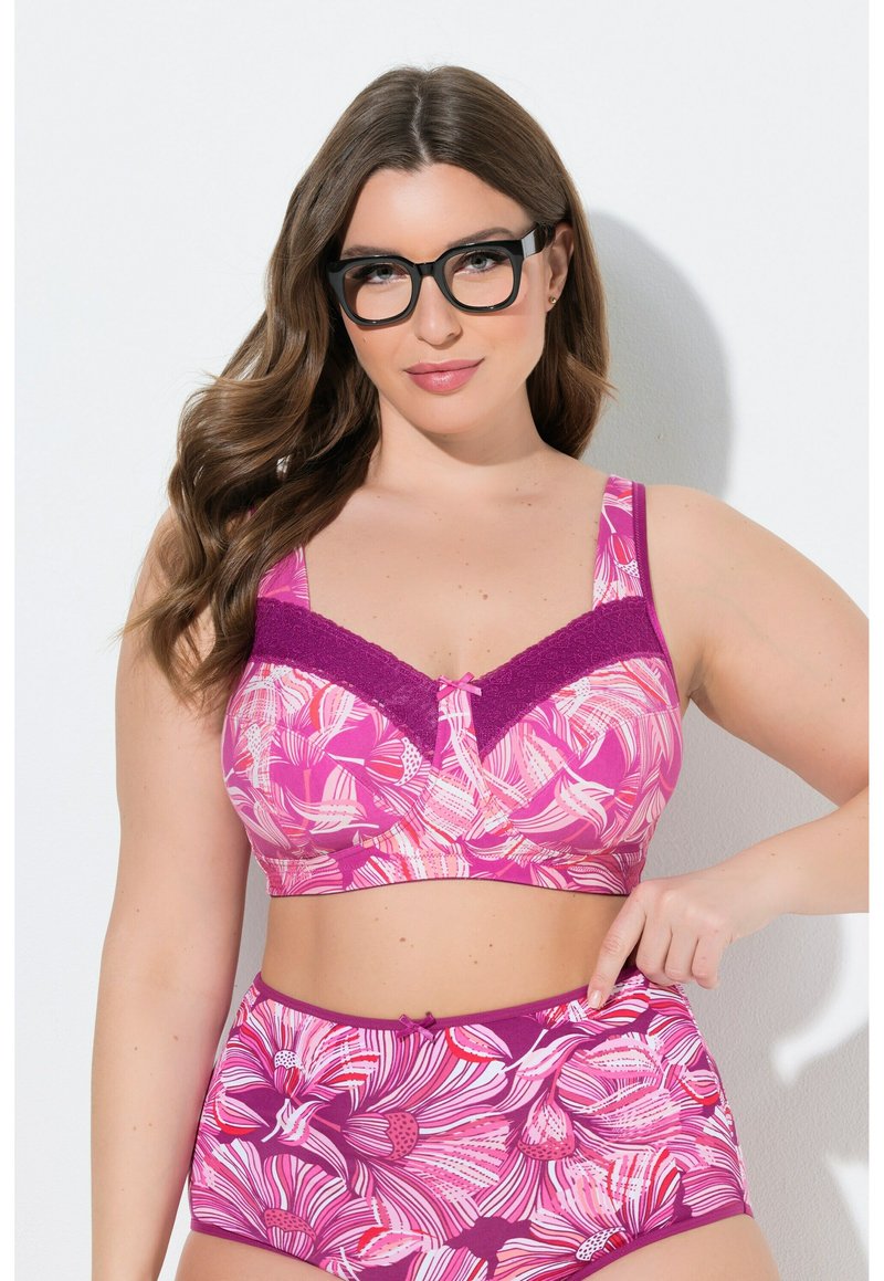 Donna con lunghi capelli castani e occhiali neri indossa un reggiseno rosa con fiori e biancheria intima a vita alta coordinata, indicando il lato della vita.