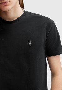 T-shirt nera in cotone con un piccolo design di teschio di capra ricamato sul petto. Collo a giro e maniche corte, texture liscia.